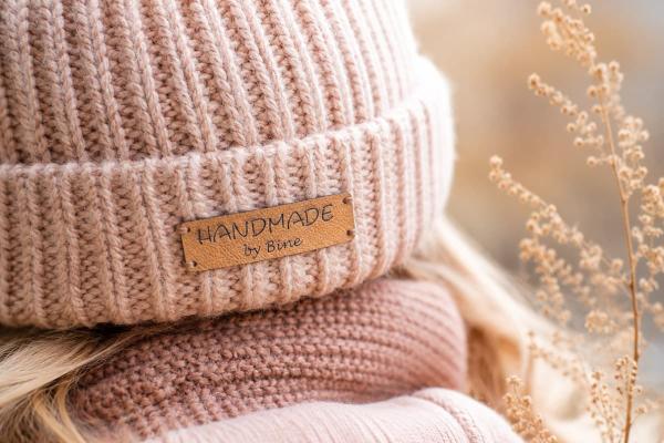 Etiqueta rectangular de polipiel marrón claro «Handmade by Bine» en gorro rosa de punto.